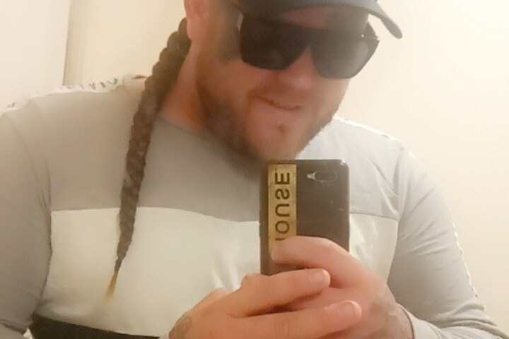 A bearded man with a plaited mullet, wearing sunglasses and a cap while taking a selfie.