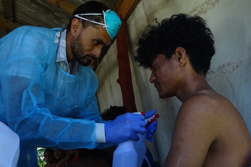 A man with a blue medical face mask on his forehead and blue scrubs sprays a young person with a bottle of water.