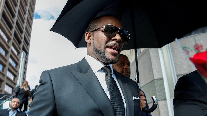 R Kelly walking outside of a courthouse with a black umbrella held over him. He's wearing a suit and sunglasses.