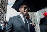 R Kelly walking outside of a courthouse with a black umbrella held over him. He's wearing a suit and sunglasses.