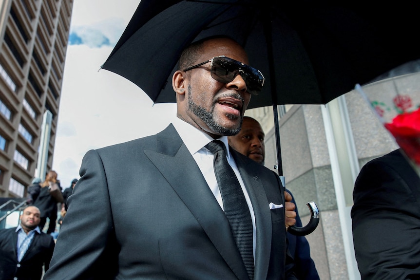 R Kelly walking outside of a courthouse with a black umbrella held over him. He's wearing a suit and sunglasses.