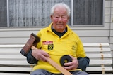 An elderly man sits on a park bench holding a mallet and wheel.