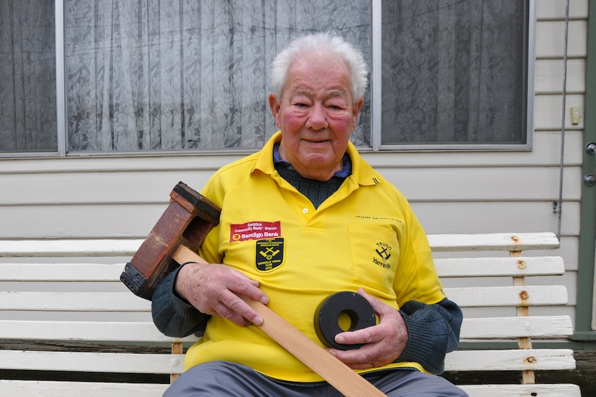 An elderly man sits on a park bench holding a mallet and wheel.