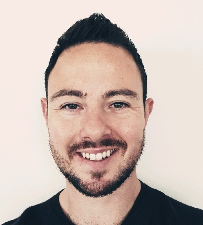 Portrait of a young man with facial hair and brown hair. He is smiling at the cmaera. 