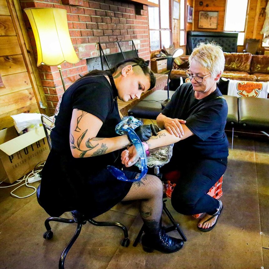 A middle-aged woman receives a tattoo from a tattooed woman