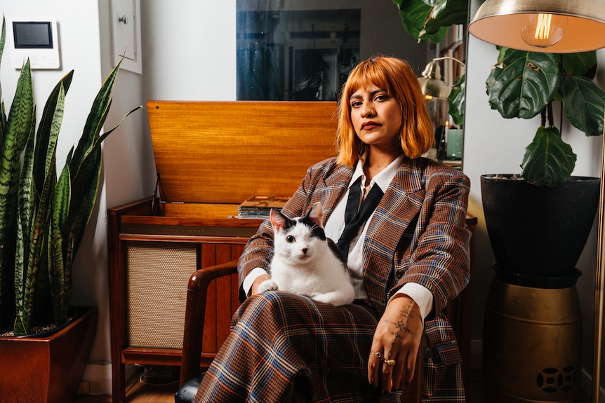 Charlie sits in a chair, in a chequered suit and tie, posing with a white and black cat on her lap