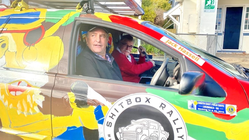 Two men sit a brightly coloured car with an emblem on the door that reads "Shitbox Rally".