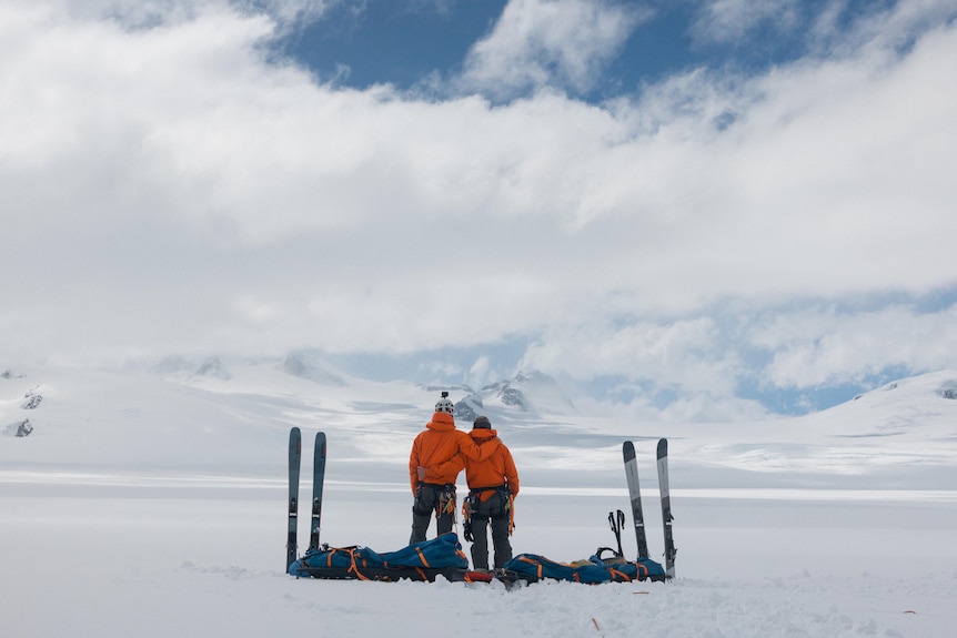 Two men stand with their arms around each other in the snow, their backs to the camera. Their ski gear is next to them.