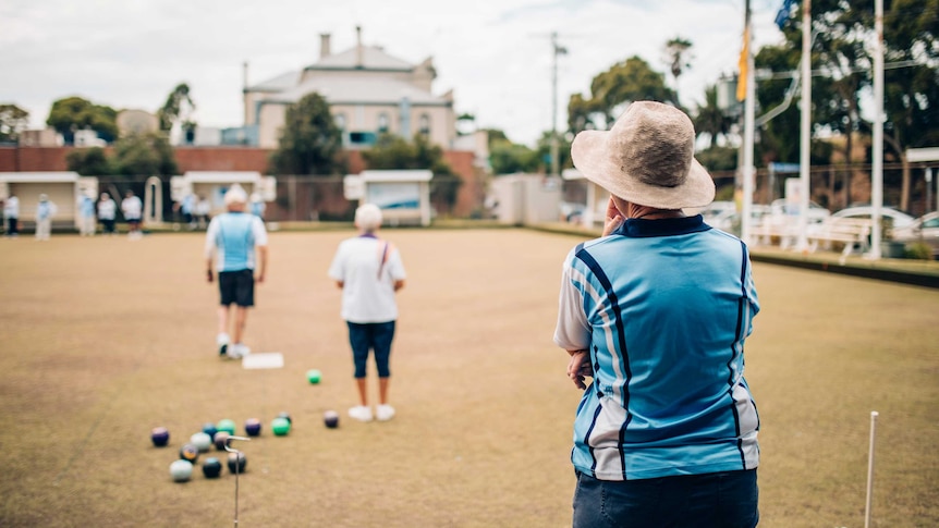 A woman in a blue shirt and hat watches a game of lawn bowls from the sideline.