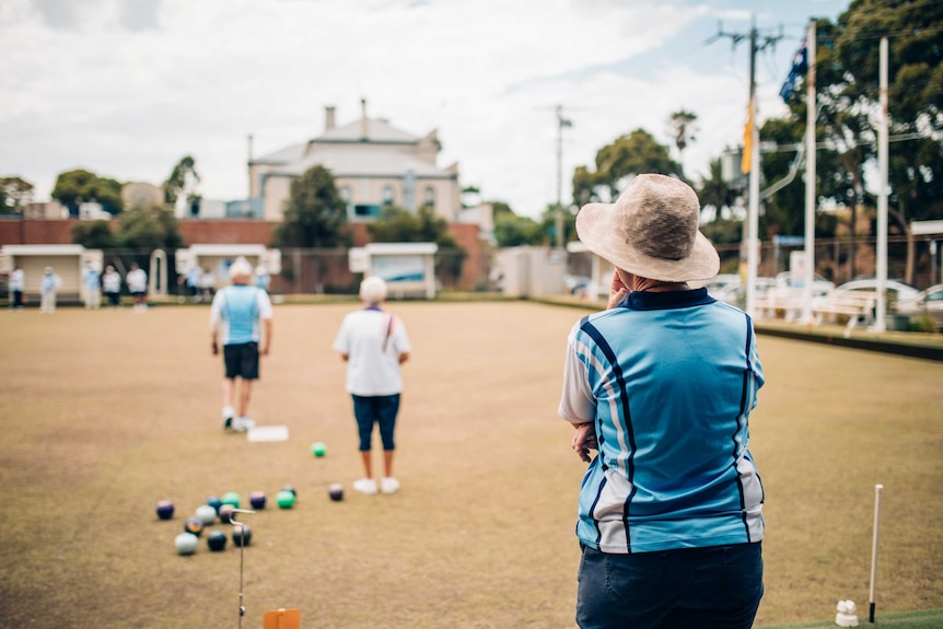 A woman in a blue shirt and hat watches a game of lawn bowls from the sideline.