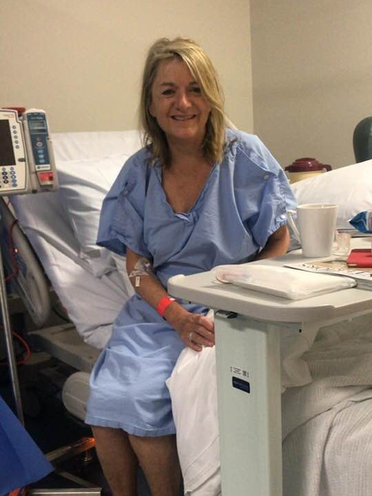 A woman sits on a hospital bed wearing a light blue hospital gown.