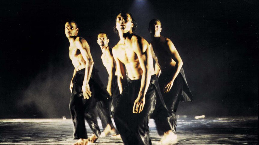 Four male dancers in long black pants and no shirt, dance in tight formation on dark stage.