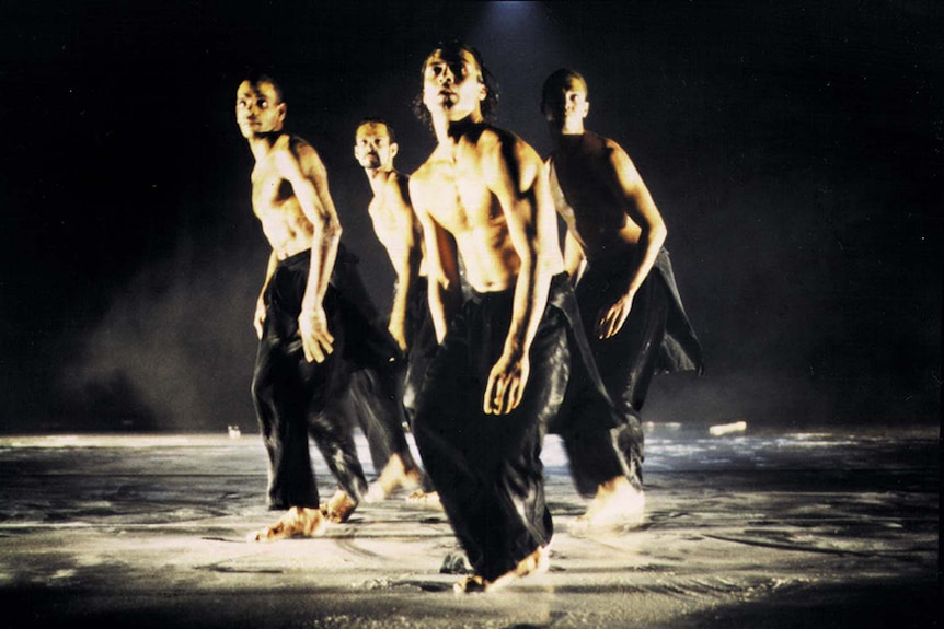 Four male dancers in long black pants and no shirt, dance in tight formation on dark stage.