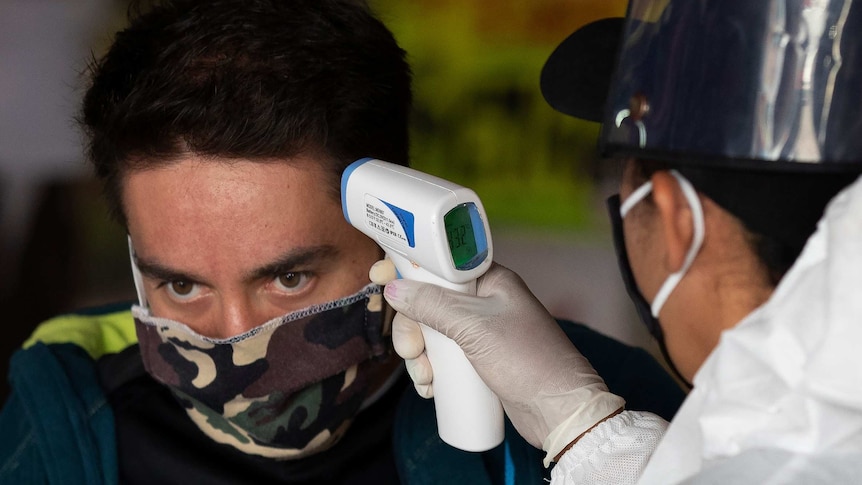 a man wearing a mask has his temperature tested with a device by a person wearing protective equipment