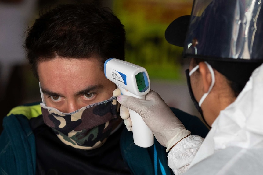 a man wearing a mask has his temperature tested with a device by a person wearing protective equipment