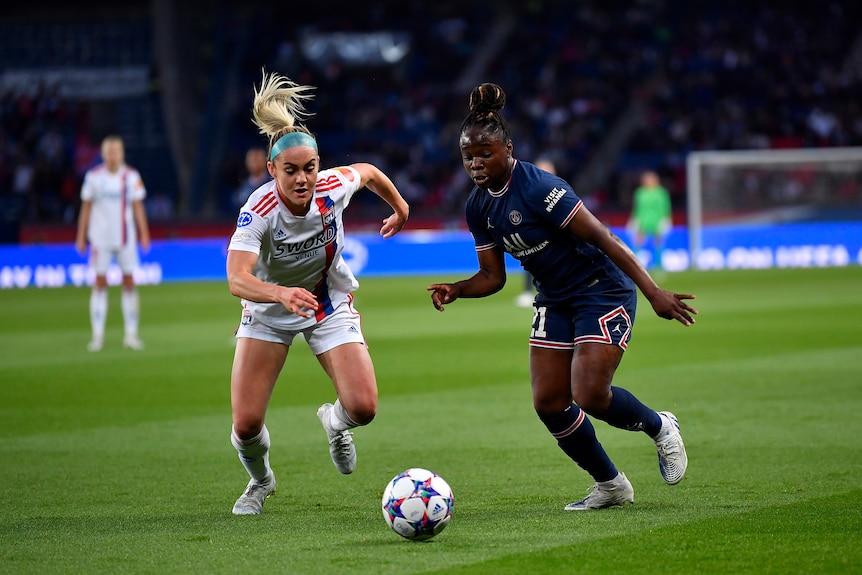 Two female soccer players, one wearing white and one wearing dark blue, compete for the ball during a match