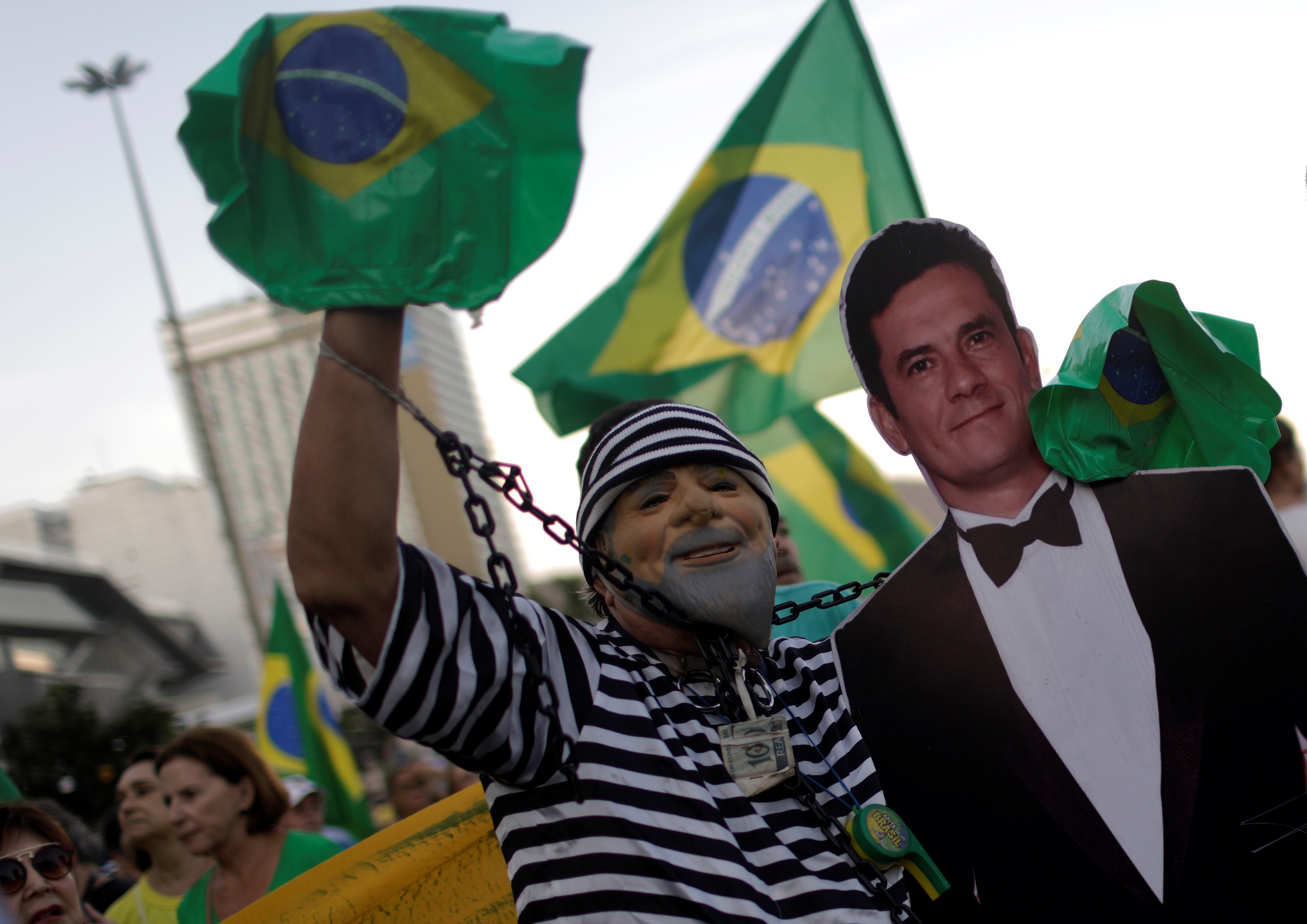 A man wearing a prisoner costume of black and white stripes, and a Lula mask, stands with a cardboard cutout