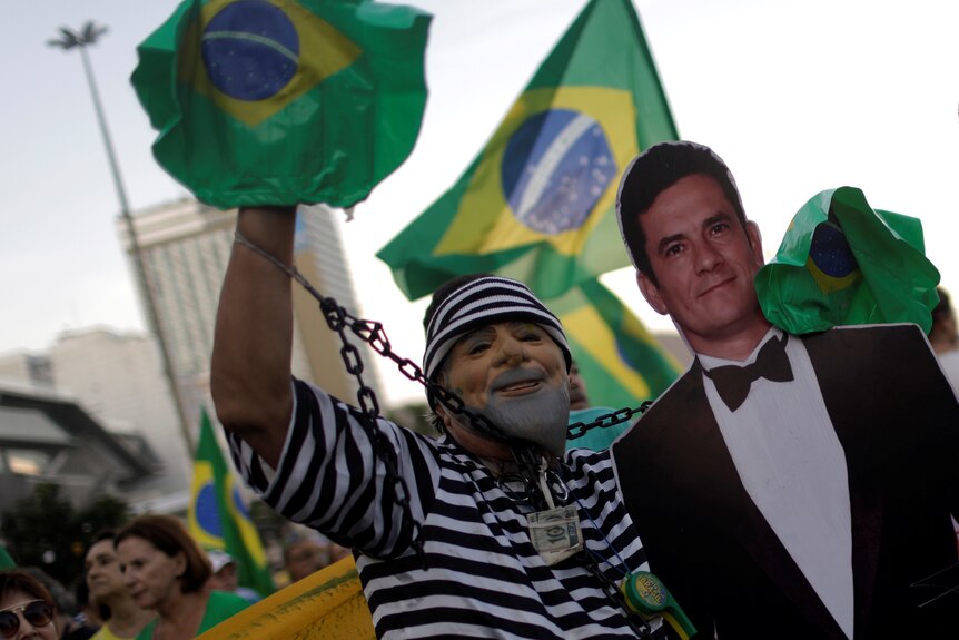 A man wearing a prisoner costume of black and white stripes, and a Lula mask, stands with a cardboard cutout