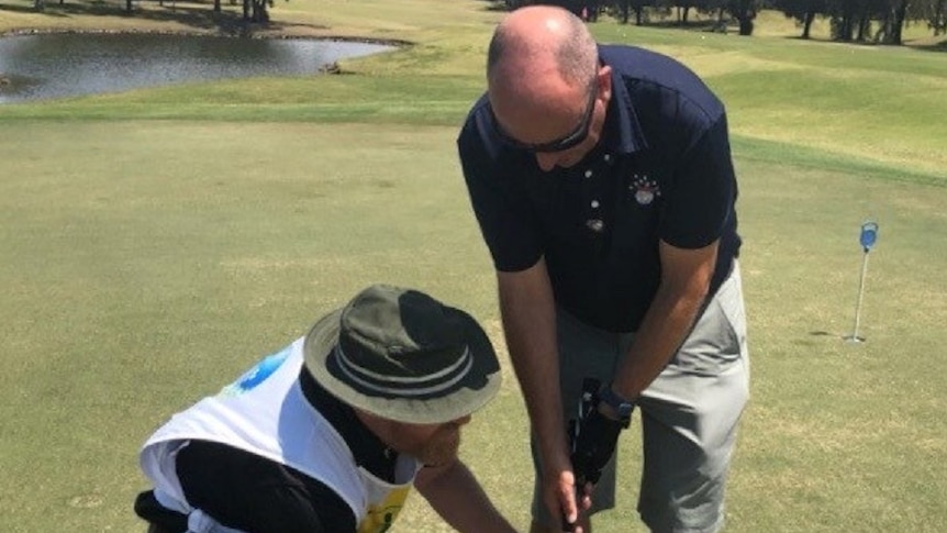 A visually impaired man bends over holding a golf putter, while another man sets the ball near the hole.