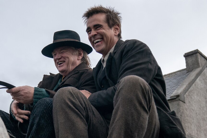 Brendan Gleeson and Colin Farrell in 20s-style dress sit smiling brightly in a wagon. 