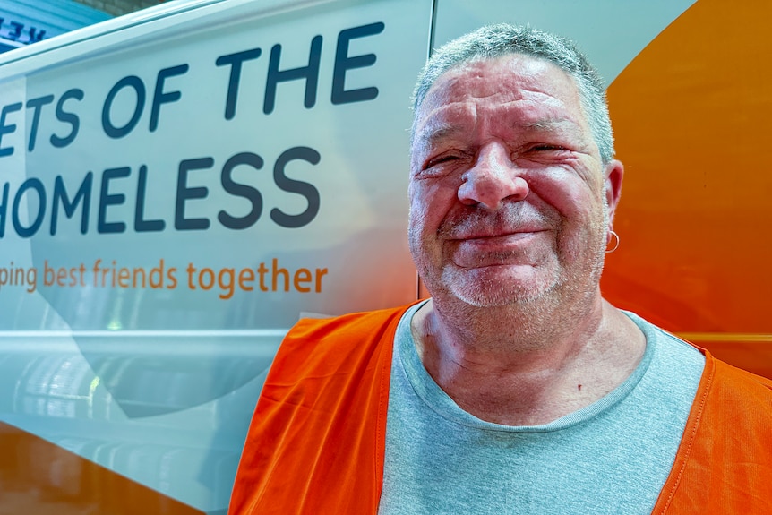 A man wears a grey t-shirt and high-vis orange vest, standing in front of a white and orange van. 