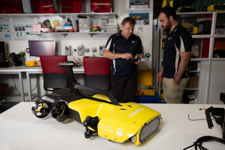 A yellow robot on a table with two men standing behind.