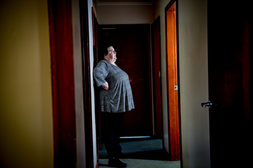 Shelly debout dans le couloir de sa maison, les mains sur les hanches, baignée de lumière naturelle, regardant par la fenêtre.