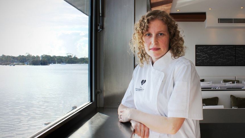 A woman in a white chef's jacket stands in a restaurant next to a river.