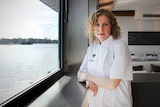 A woman in a white chef's jacket stands in a restaurant next to a river.