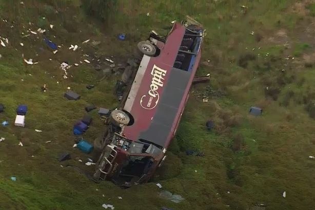 A bus lying on its side at the bottom of a hill with debris strewn for metres.