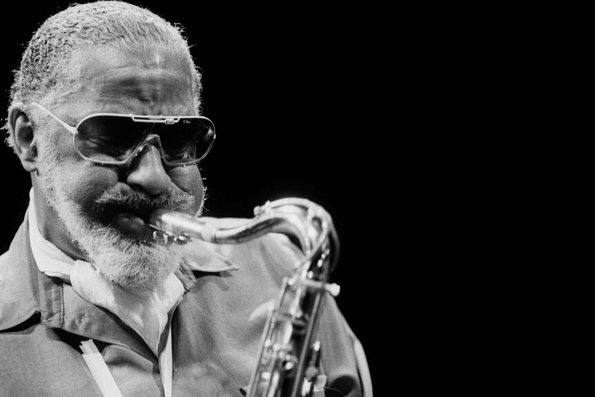 Black and white photo of man with sunglasses, beard and moustache, blows hard into the mouthpiece of a saxophone he's holding.