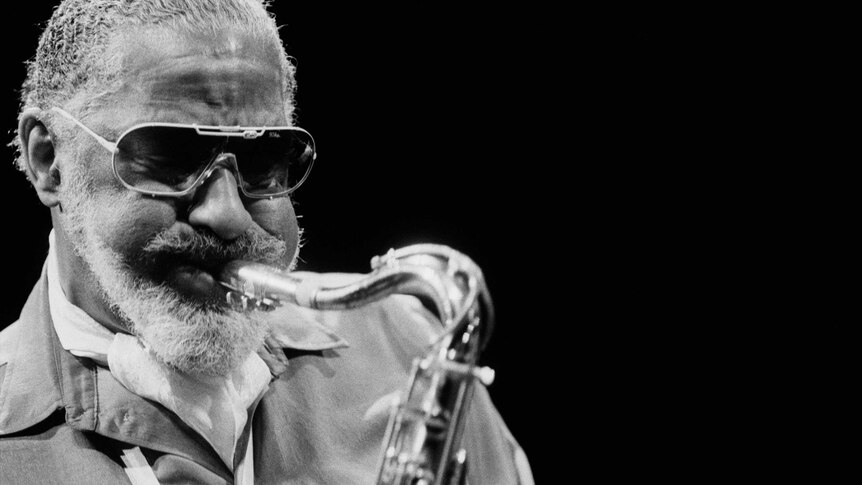Black and white photo of man with sunglasses, beard and moustache, blows hard into the mouthpiece of a saxophone he's holding.