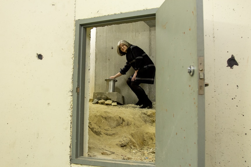 A view through a door of a woman placing a metal cylinder atop a concrete block that sits on dirt and rubble.
