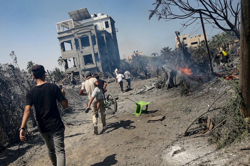 People walk toward a bombed-out building past debris and a small fire