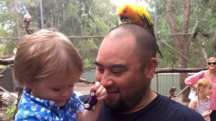 Jacob Lee Moore with a yellow parrot on his head, holding his young grandson who holds a toy car.