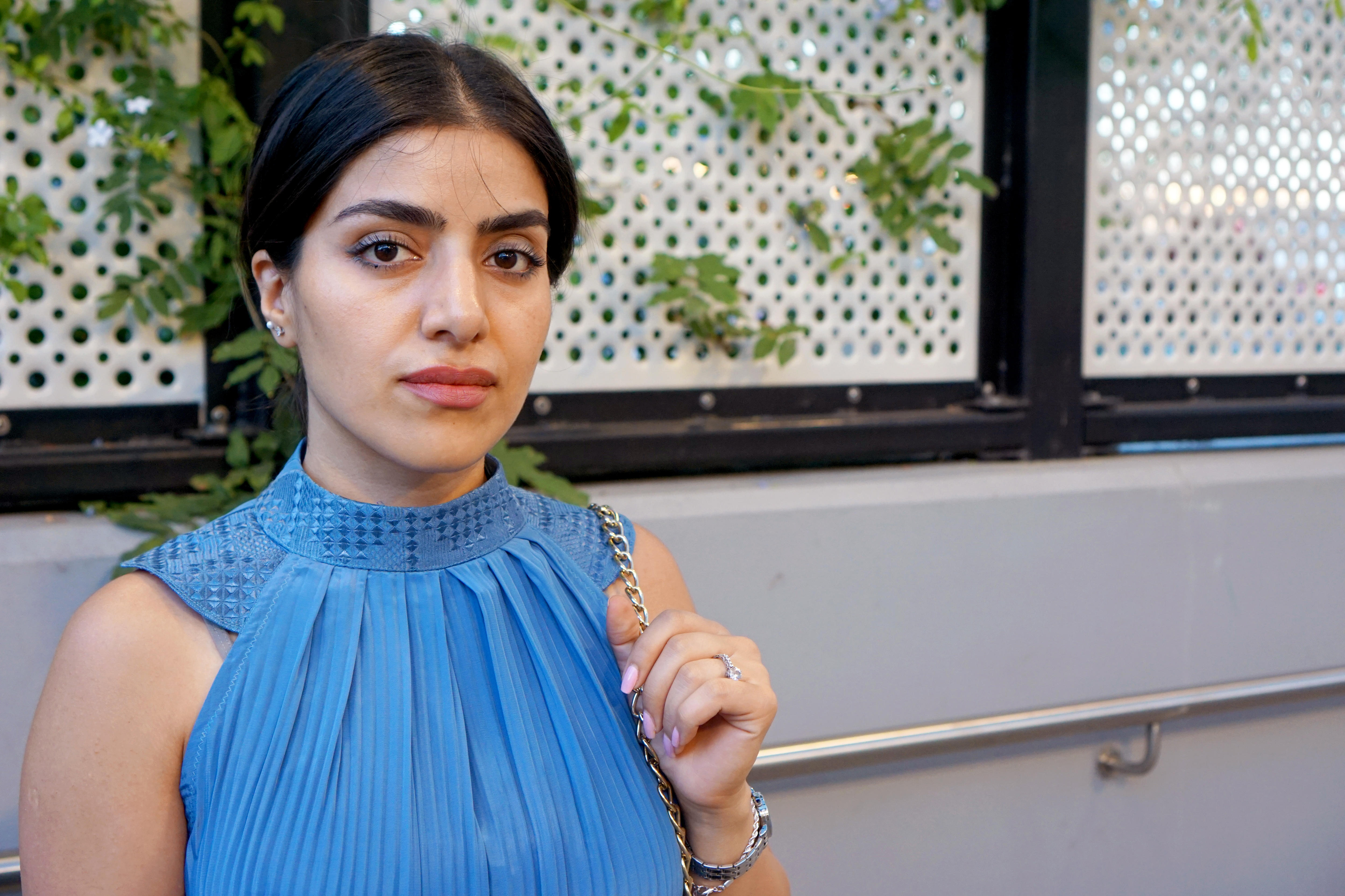 A young Afghan woman wearing a blue high-necked, sleeveless dress poses for a photo.