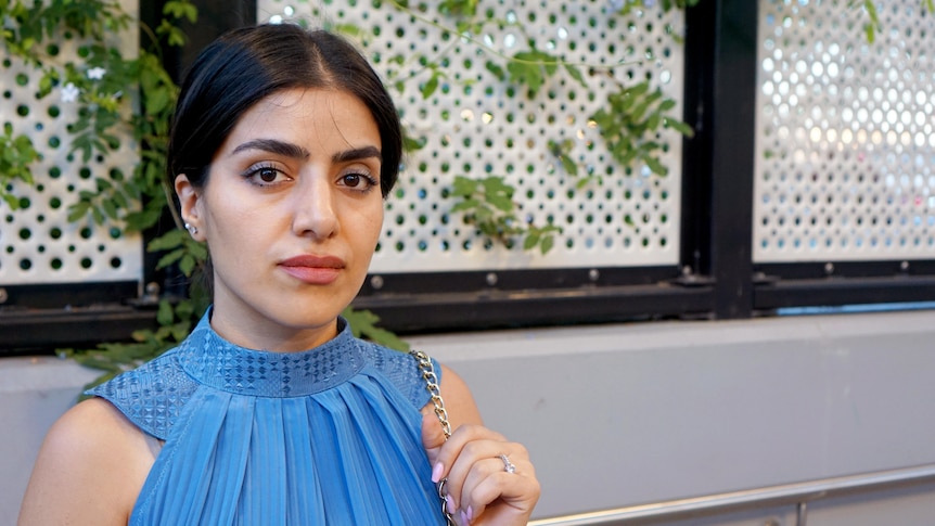 A young Afghan woman wearing a blue high-necked, sleeveless dress poses for a photo.