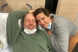 Anthony Ellison smiles while lying in a hospital bed. His wife Clair is beside him.