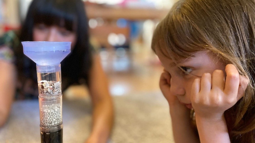 A girl looks at a science experiment as her mother observes in the background.