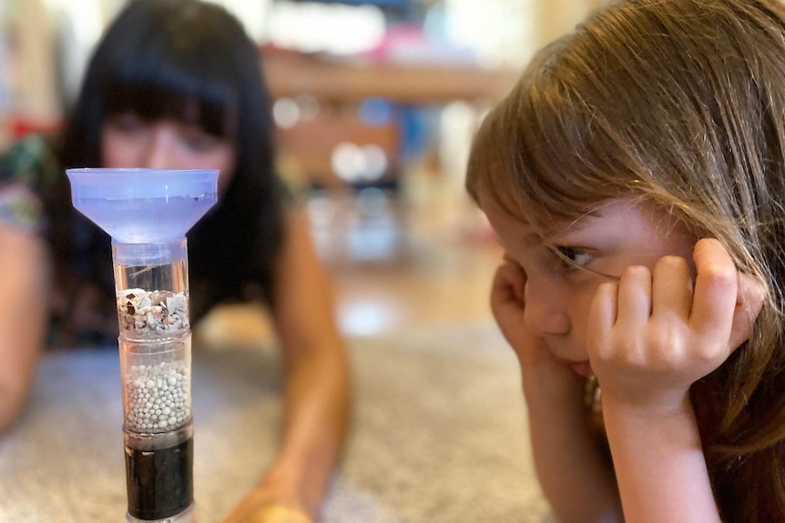 A girl looks at a science experiment as her mother observes in the background.