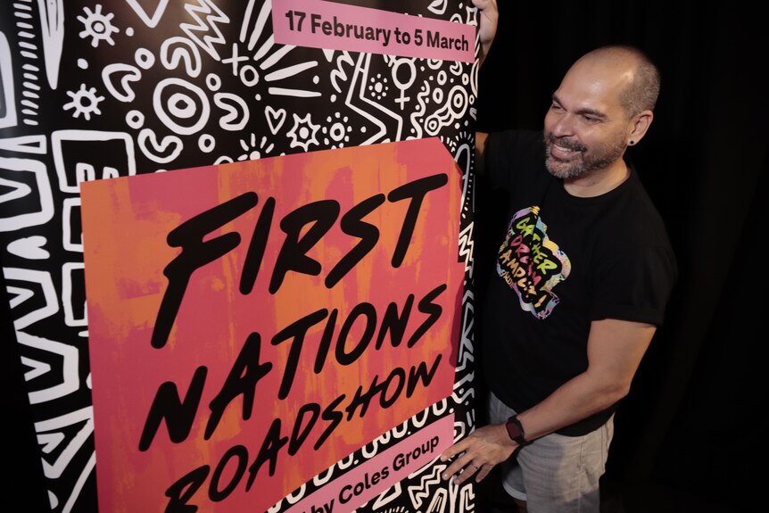 A man stands next to a sign advertising the First Nations Roadshow