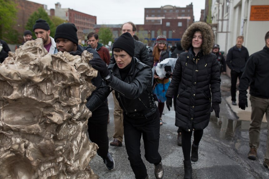 The artist and others determinedly push a lumpy sculpture.