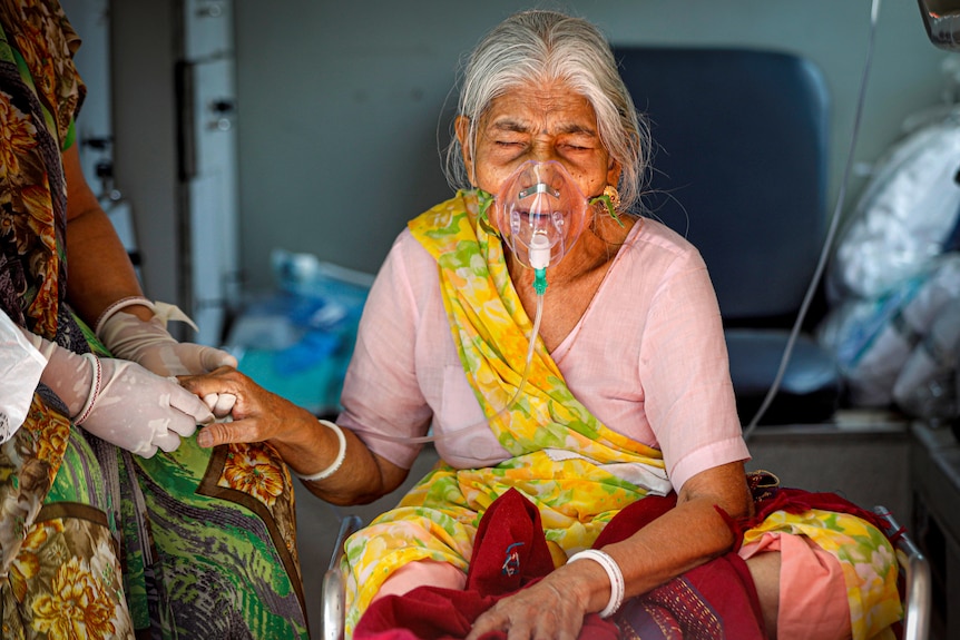 An older Indian woman in a yellow, red and pink sari with an oxygen mask on her face, while another woman holds her hand