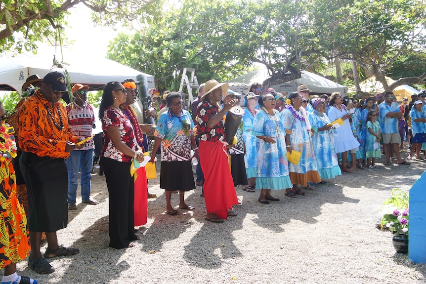 A group of people lined up along a path singing. One man has an island drum.