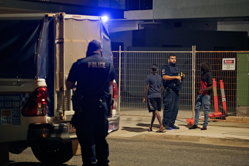 Two police officers and two people stand near a police wagon at night. 