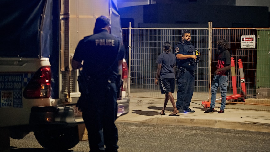 Two police officers and two people stand near a police wagon at night. 