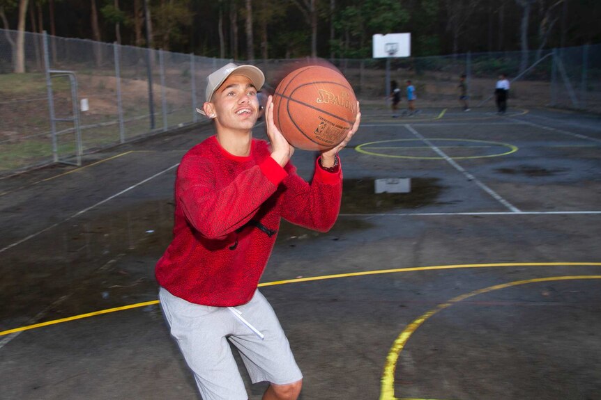 A boy in a red shirt and white cap shooting a basketball at a hoop.