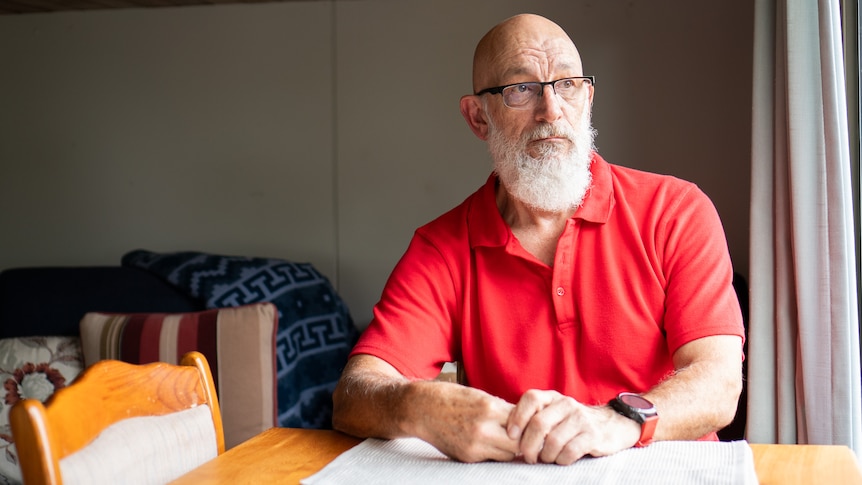 A bald man with a white beard and red top sits at a table.