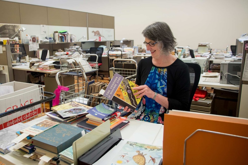 A woman sits and holds a book, surrounded by stacks of other books.