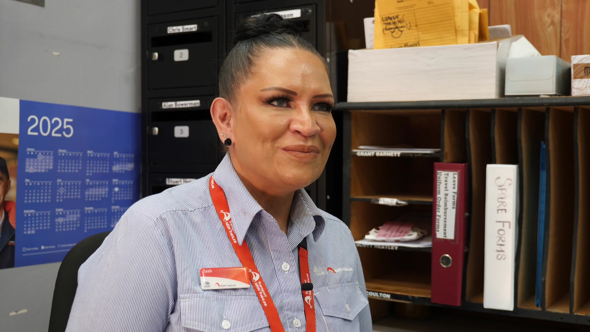 Uma mulher com o cabelo preso e um cordão laranja sorri. Ela está em um escritório.